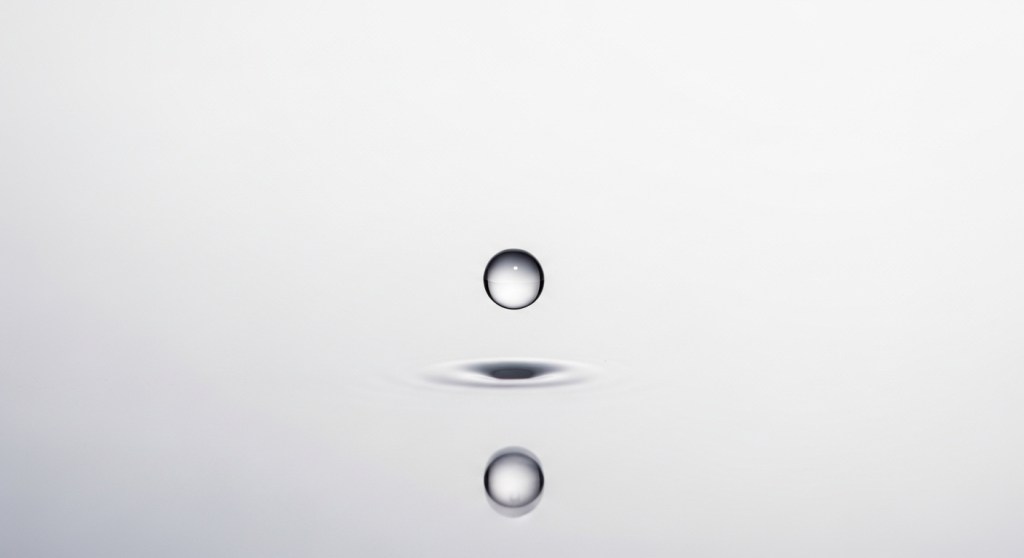 Single, perfect clear liquid droplet suspended above a calm, reflective grey surface.